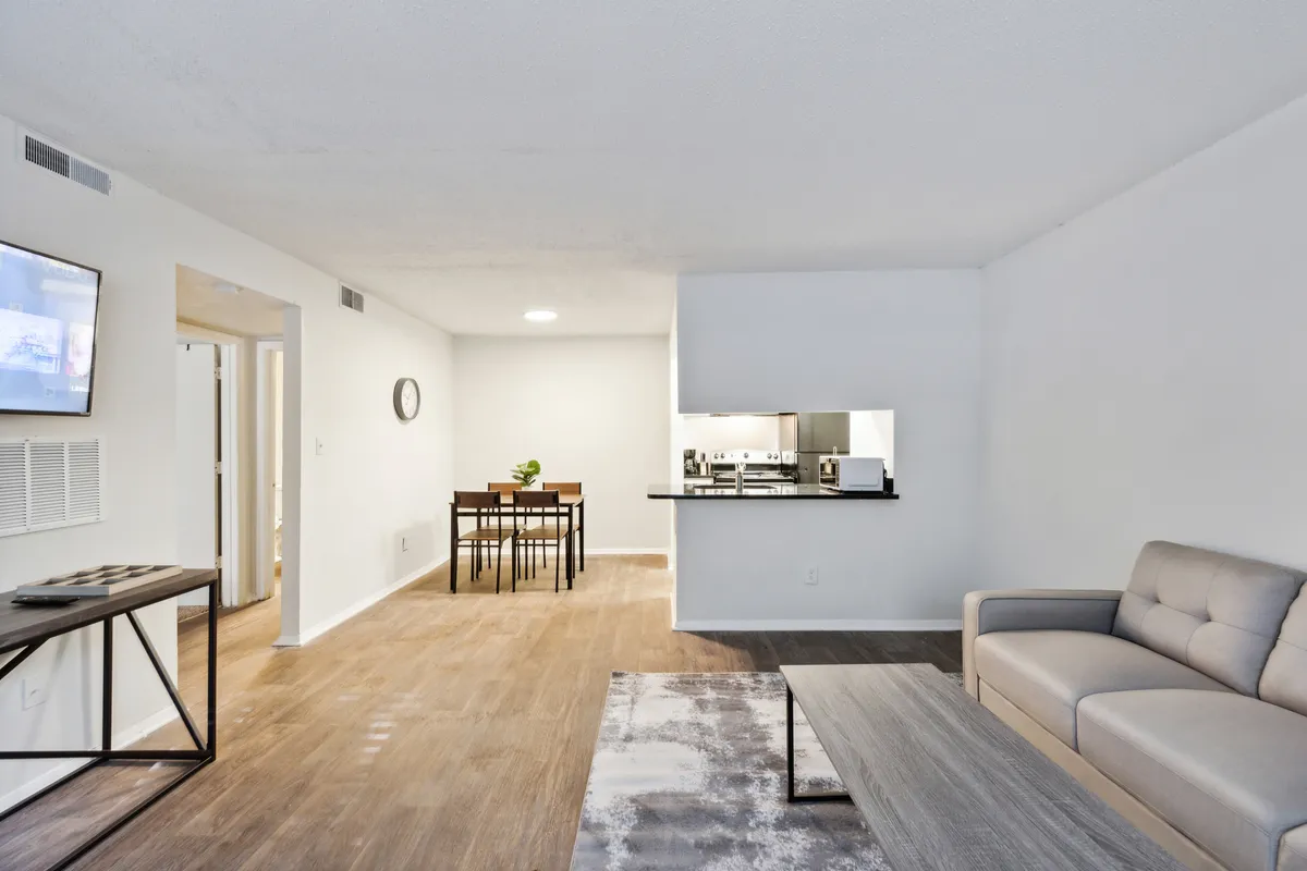 This image showcases a contemporary living area with a comfortable sofa, dining table, TV, and a view into the kitchen. The open-plan layout is inviting and modern, suggesting both relaxation and socializing. Large windows in the space hint at a connection to the outside, reinforcing the feeling of being in an urban, vibrant environment.
