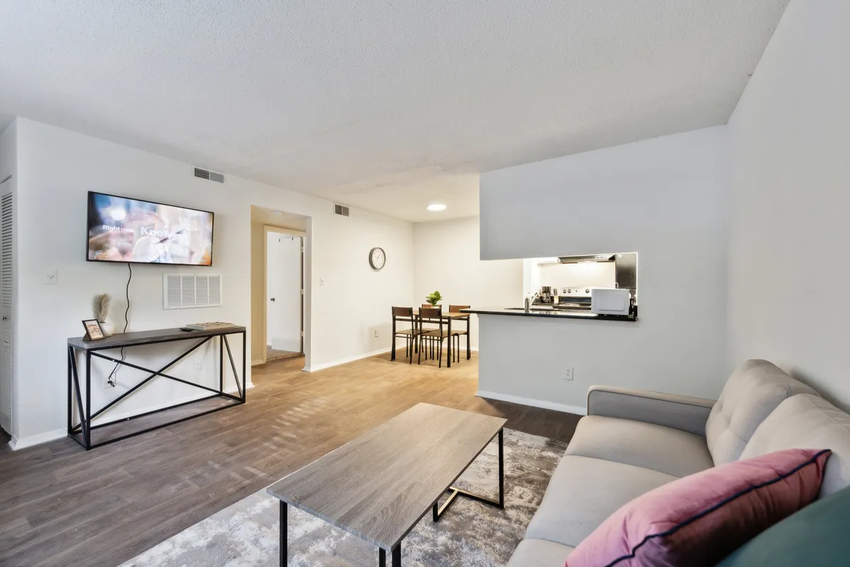 This image showcases a bright, comfortable living area with a plush sofa, coffee table, and a TV in clear view. The open layout seamlessly extends to the adjacent kitchen and dining table, depicting a connected, multi-functional space ideal for both relaxation and entertaining. The scene is alive with natural light, emphasizing the room’s inviting atmosphere.