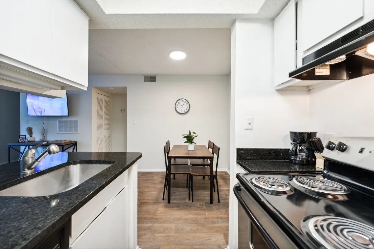 A kitchen and dining space featuring a clean countertop, dining table set for a casual meal, modern appliances, and a wall clock. The layout is open and practical, encouraging easy transitions from meal prep to socializing or working remotely.