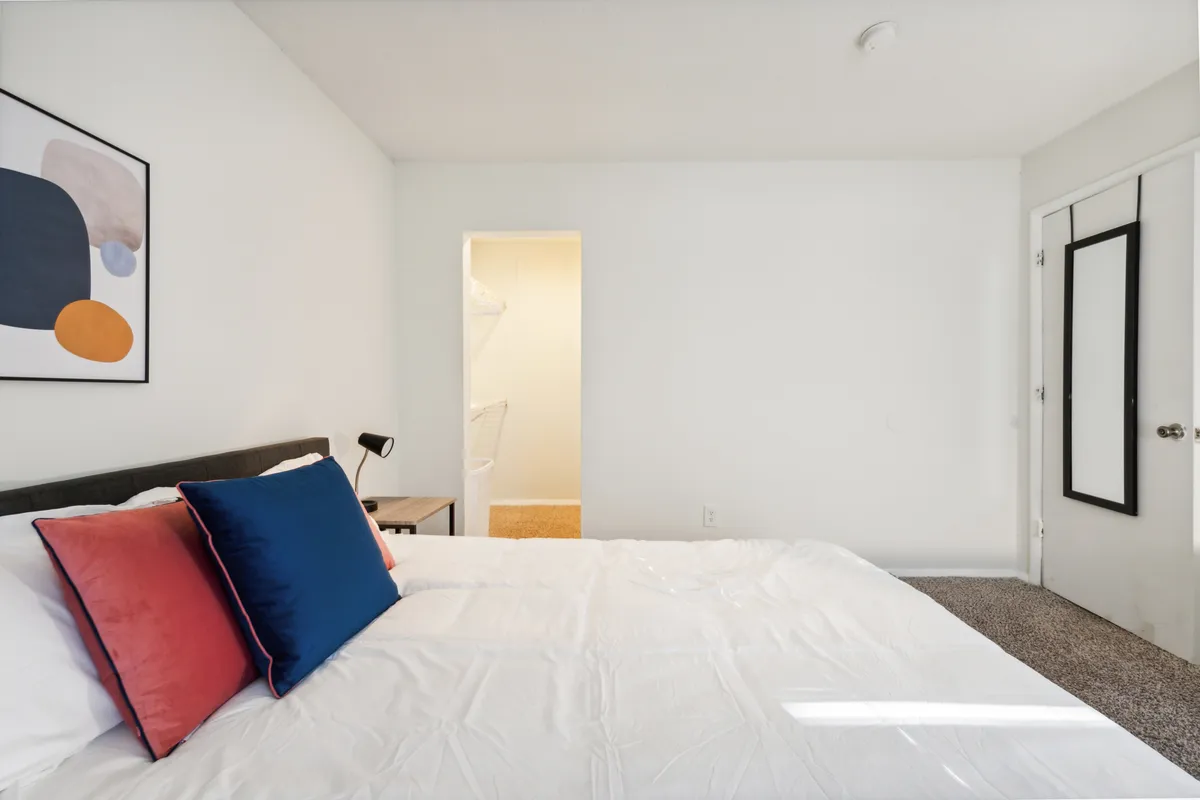 A cozy bedroom with a plush bed topped with decorative pillows, soft bedding, and warm lighting from a bedside lamp. Artwork adorns the wall above the bed, creating a serene atmosphere perfect for relaxation or quiet moments alone.