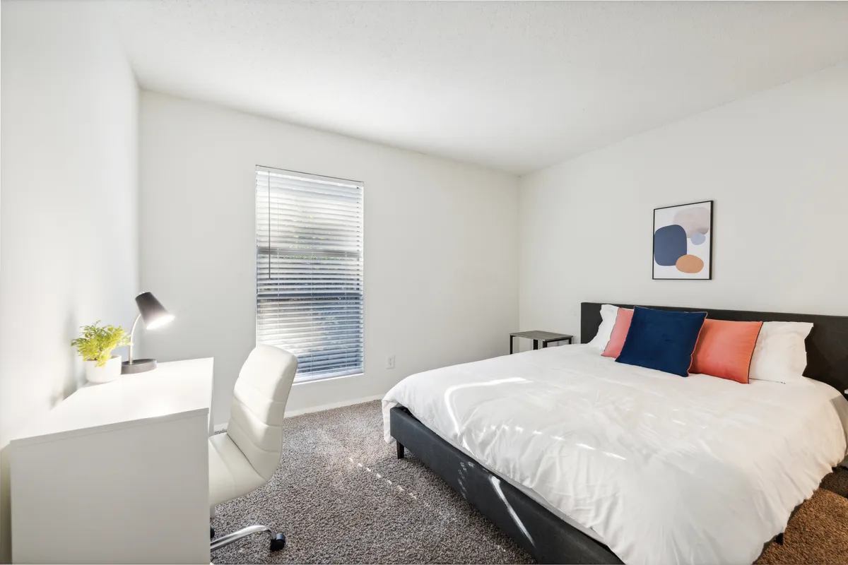The image features a serene, sunlit bedroom with a plush bed, a modern desk positioned near a large window, and tasteful artwork adorning the walls. Natural light floods the space, creating an inviting atmosphere perfect for both relaxation and productivity.